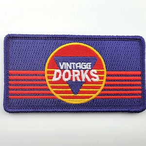 May include: A rectangular embroidered patch with a dark blue background and a central yellow circle containing a purple triangle. The text VINTAGE DORKS is stitched in white across the center, with four horizontal red lines extending outward.
