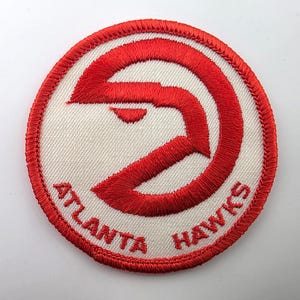 May include: A circular embroidered patch featuring the red Atlanta Hawks logo on a white background. The design includes a stylized red hawk emblem centered above the red text Atlanta Hawks, all enclosed within a thick red stitched border.