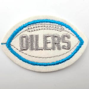 May include: An oval-shaped embroidered patch on a white felt base. The patch features a bright blue stitched border and silver metallic thread detailing. The word OILERS is embroidered in silver thread across the center of the football-shaped design.