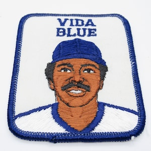 May include: A rectangular embroidered patch featuring a portrait of a person wearing a blue baseball cap. The text VIDA BLUE is stitched in blue above the figure. The patch has a white background and a thick blue stitched border edge.