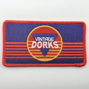 May include: A rectangular embroidered patch with a purple background and a red border. It features a central yellow circle containing a blue triangle and the white text VINTAGE DORKS. Four horizontal red stripes extend across the purple field.