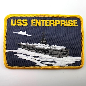 May include: A rectangular embroidered patch with a dark blue background and a yellow border. The text USS ENTERPRISE is stitched in yellow at the top. Below, a grey aircraft carrier is depicted sailing on white embroidered water waves.