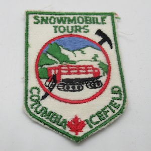 May include: A shield-shaped embroidered patch featuring the text SNOWMOBILE TOURS and COLUMBIA ICEFIELD. The design includes a red snowcat vehicle on a blue and green mountain landscape, a black pickaxe icon, and a red maple leaf at the bottom.