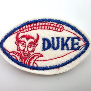 May include: An oval-shaped embroidered patch with a white felt base. It features a red stitched graphic of a devil character on the left and the blue embroidered text DUKE on the right. The entire patch is bordered by a thick blue stitched edge.