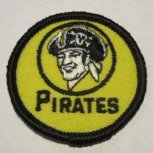 May include: A circular embroidered patch featuring a yellow background with a black border. In the center, a black and white illustration of a pirate wearing a tricorn hat is depicted above the word PIRATES embroidered in black capital letters.