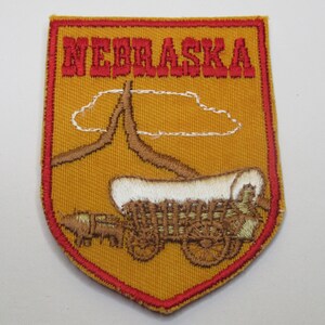 May include: A shield-shaped embroidered patch with a mustard yellow background and a red border. The text NEBRASKA is stitched in red at the top. Below, a brown covered wagon pulled by a horse is depicted with a white cloud outline above.