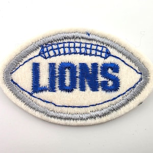 May include: An oval-shaped embroidered patch featuring a cream-colored felt base with a silver-grey stitched border. The center displays the word LIONS in bold blue thread, with a blue stitched graphic resembling a football lace above the text.