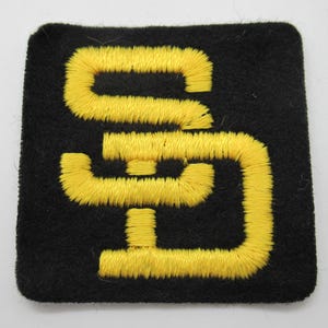 May include: A square black fabric patch featuring the letters SD embroidered in bright yellow thread. The letters are stylized and interconnected, with the S positioned above the D, creating a bold contrast against the dark background.