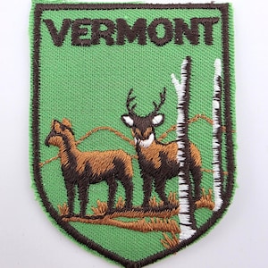 May include: A shield-shaped embroidered patch with a light green background and a dark brown border. The text VERMONT is stitched in dark brown at the top. It features two brown deer standing near two white and black birch tree trunks.