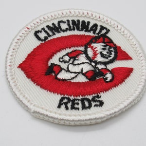 May include: A circular embroidered patch featuring a white background with a red letter C. Inside the C is a cartoon character wearing a baseball cap and uniform. The text CINCINNATI is arched above the character, and the word REDS is below it.