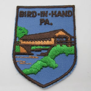 May include: A shield-shaped embroidered patch with a light blue background and a dark brown border. It features a brown covered bridge over a stream with green foliage and the text BIRD-IN-HAND PA. stitched in dark brown thread at the top.