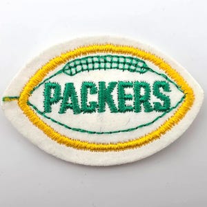 May include: An oval-shaped embroidered patch on a white felt background. The design features the word PACKERS in green thread, surrounded by a green stitched football outline and a thick yellow border, mimicking the shape of a football.