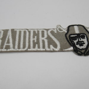 May include: A rectangular embroidered patch featuring the word RAIDERS in white capital letters on a grey felt background. To the right of the text is an embroidered graphic of a person wearing a helmet with two crossed swords behind it.