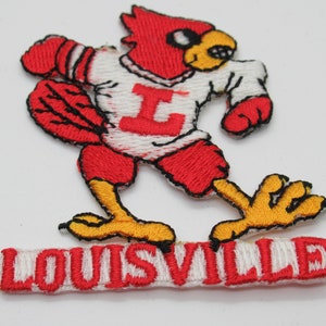 May include: An embroidered patch featuring a red cardinal mascot wearing a white jersey with a red letter I on the chest. The cardinal has yellow feet and a yellow beak. Below the mascot, a white rectangular patch displays the word LOUISVILLE in red.