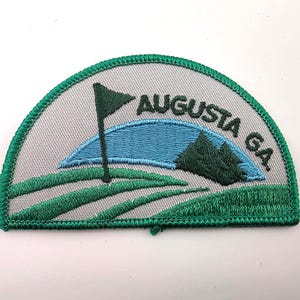 May include: A semi-circular embroidered patch featuring a golf course scene with a green flag, rolling green hills, a blue sky arc, and dark green trees. The text AUGUSTA GA. is embroidered in dark green thread against a light gray background.