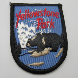 May include: A shield-shaped embroidered patch with a light blue background and a black stitched border. It features a black bear, a white geyser plume, and the red embroidered text Yellowstone Park. The design includes dark green trees.