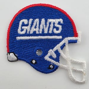 May include: An embroidered patch shaped like a football helmet in blue, red, and white. The helmet features the word GIANTS in white block letters across the side, with a white face mask and a red stripe along the top edge of the helmet.