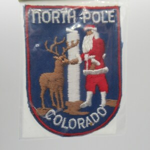 May include: A rectangular embroidered patch with a blue background and a red border. It features the text NORTH POLE at the top and COLORADO at the bottom. The design shows a brown reindeer and Santa Claus standing on either side of a white pole.