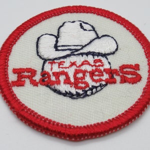 May include: A circular embroidered patch featuring a white background with a red stitched border. The design includes a white cowboy hat sitting atop a baseball, with the red embroidered text Texas Rangers displayed across the center of the ball.
