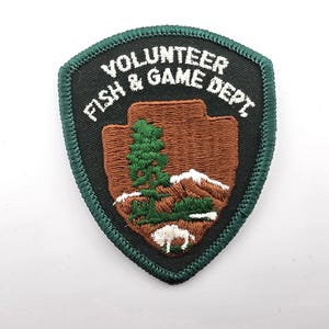 May include: A shield-shaped embroidered patch with a black background and a dark green border. The text VOLUNTEER FISH & GAME DEPT is stitched in white. The center features a brown arrowhead shape with a green pine tree, mountains, and a white bison.