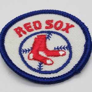 May include: A circular embroidered patch featuring the text RED SOX in red thread above a graphic of two red socks arranged in a cross pattern, all set against a white background with a blue stitched border and a blue baseball stitch design.