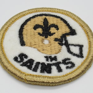 May include: A circular embroidered patch featuring a gold football helmet with a black fleur-de-lis logo. The word SAINTS is stitched in black thread below the helmet. The patch has a white background and a thick, metallic gold stitched border.