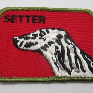 May include: A rectangular embroidered patch with a red background and an olive green stitched border. The patch features a black and white profile illustration of a dog head and the word SETTER embroidered in black text in the upper left corner.