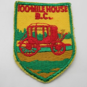 May include: A shield-shaped embroidered patch featuring a red stagecoach on a yellow background with green forest details. The text 100 MILE HOUSE B.C. is stitched in red at the top. The patch has a green border and a yellow outer edge finish.
