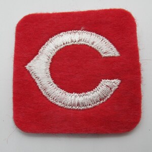 May include: A square red felt patch featuring a white embroidered letter C in the center. The letter C has a pointed serif on the left side and a rounded shape, with dense white stitching creating a textured appearance against the red fabric.