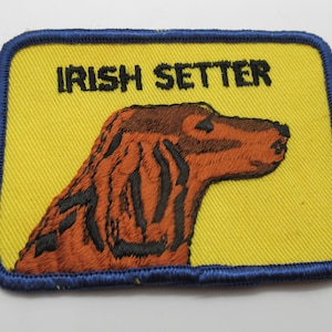 May include: A rectangular embroidered patch with a yellow background and a blue border. It features an embroidered profile of a brown Irish Setter dog and the black text IRISH SETTER printed above the dog's head on the yellow fabric surface.