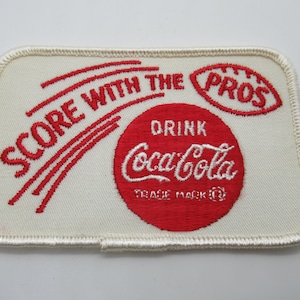 May include: A rectangular embroidered patch with an off-white background and a cream-colored stitched border. The patch features red embroidery that reads "SCORE WITH THE PROS" above a red circle containing the white text "DRINK Coca-Cola".