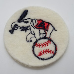 May include: A circular off-white felt patch featuring an embroidered design of an elephant standing on a baseball. The elephant is outlined in dark blue thread, wears a red blanket with a dark blue letter A, and holds a dark blue baseball bat.