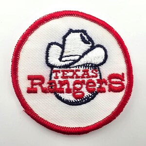May include: A circular embroidered patch featuring a white baseball with navy blue stitching and a white cowboy hat on top. The text Texas Rangers is embroidered in red across the center of the baseball. The patch has a thick red stitched border.
