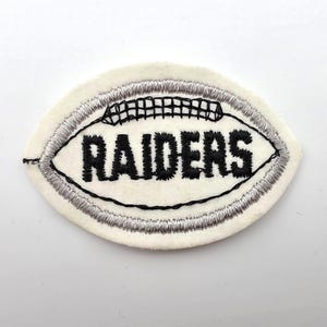 May include: An oval-shaped embroidered patch featuring a white background with a silver stitched border. The center displays the word RAIDERS in bold black lettering, with black stitching detailing the laces of a football shape.