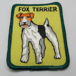 May include: A rectangular embroidered patch with a yellow background and a dark green border. The patch features a stitched illustration of a Fox Terrier dog with white, black, and orange fur. The text FOX TERRIER is embroidered in black at the top.