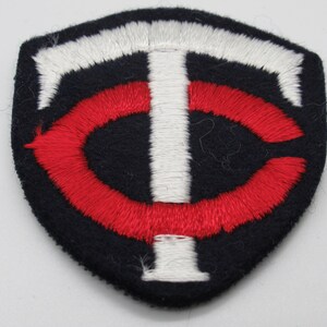 May include: A shield-shaped embroidered patch featuring a dark navy blue background with a white letter T and a red letter C intertwined in the center. The design is stitched with thick thread, creating a textured surface on the fabric base.