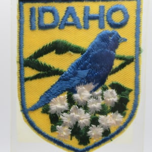 May include: A shield-shaped embroidered patch with a yellow background and a blue border. The text IDAHO is stitched in blue at the top. It features a blue bird perched on white flowers with green leaves, set against a dark green mountain silhouette.