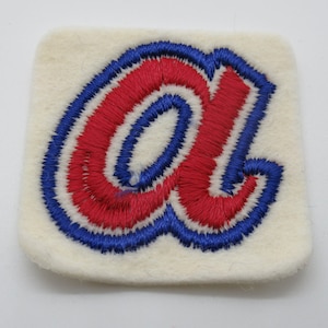 May include: A square-shaped off-white felt patch featuring a red embroidered lowercase letter a with a thick blue border. The embroidery is dense and textured, creating a raised effect against the flat, light-colored felt background material.