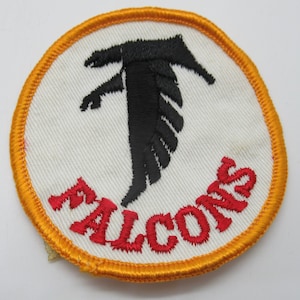May include: A circular embroidered patch featuring a white background with a black falcon silhouette in the center. The word FALCONS is stitched in red thread below the bird. The entire patch is bordered by a thick, vibrant orange stitched edge.