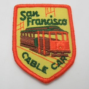 May include: A shield-shaped embroidered patch with a yellow background and an orange border. It features a brown and red cable car illustration, green embroidered text reading San Francisco and Cable Car, and a small red graphic symbol.