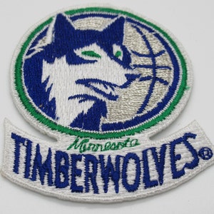May include: A circular embroidered patch featuring a blue and white wolf head silhouette layered over a basketball graphic, bordered by a green ring. Below the circle, a white banner displays the text Minnesota TIMBERWOLVES in blue and green.