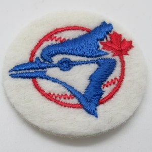 May include: A circular white felt patch featuring an embroidered blue jay bird silhouette in blue thread. The design includes a red circular border and a small red maple leaf detail. The embroidery is textured and centered on the white fabric base.