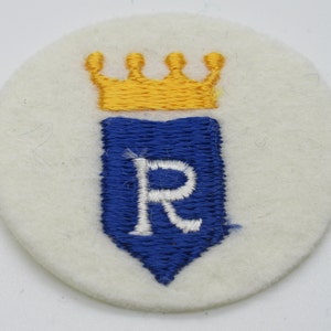 May include: A circular off-white felt patch featuring an embroidered blue shield centered with a white letter R. Above the shield is an embroidered yellow crown. The design is stitched directly onto the circular fabric base with visible thread.