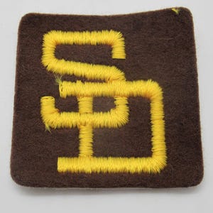 May include: A square-shaped brown felt patch featuring the yellow embroidered letters SD in a stylized, interlocking font. The embroidery has a raised, textured appearance with visible thread stitching throughout the yellow lettering.