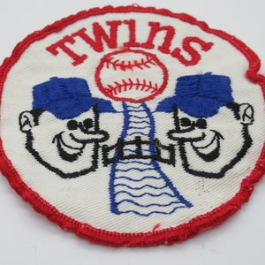 May include: A circular embroidered patch with a white background and a red stitched border. It features the word TWINS in red, a red baseball, and two cartoon faces wearing blue caps, separated by a blue bridge graphic with black outlines.