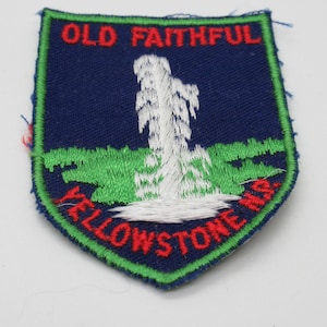 May include: A shield-shaped embroidered patch with a dark blue background and a bright green border. It features a white embroidered geyser erupting from a green grassy area. The text OLD FAITHFUL is in red at the top and YELLOWSTONE N.P. is in red at the bottom.