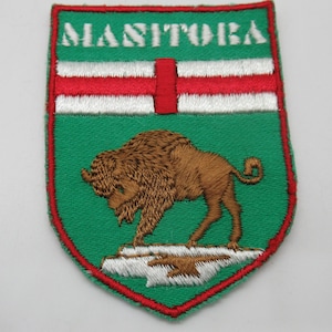 May include: A shield-shaped embroidered patch featuring a green background with a red border. The word MANITOBA is stitched in white at the top. A brown bison stands on a white rock formation above a horizontal white and red striped band.