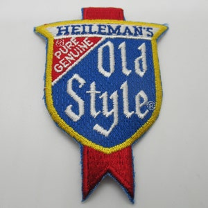 May include: A shield-shaped embroidered patch featuring a blue background with white text that reads HEILEMAN'S OLD STYLE. A red diagonal banner contains the white text PURE GENUINE. The patch is bordered by yellow stitching and has red accents.