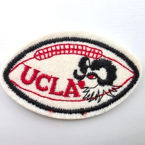 May include: An oval-shaped embroidered patch with a white felt background. It features the red text UCLA, a red stitched arch design above the text, and a black and red embroidered ram head mascot on the right side, all bordered by black stitching.