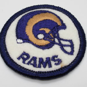May include: A circular embroidered patch featuring a blue and gold football helmet design on a white background. The word RAMS is stitched in blue thread below the helmet. The entire patch is bordered by a thick, dark blue stitched edge.